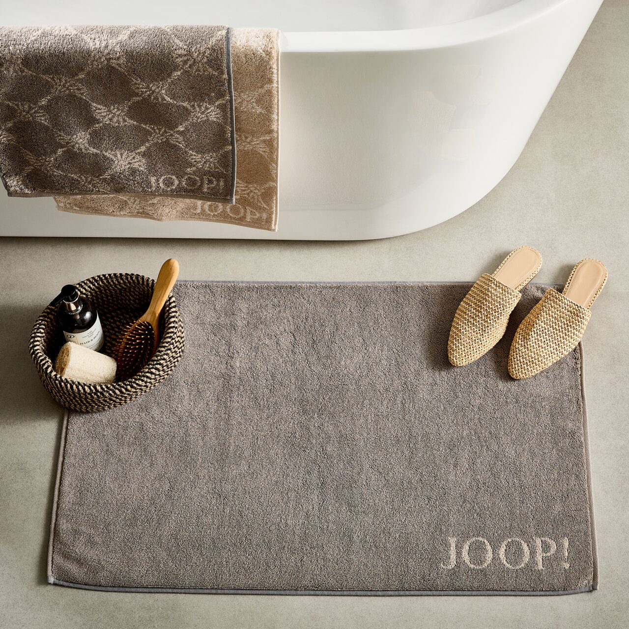 JOOP! Badematte DOUBLEFACE Graue Badematte mit 'JOOP!' Schriftzug, platziert vor einer Badewanne, umgeben von einem Korb mit Pflegeprodukten und beigen Hausschuhen, aus der Vogelperspektive.