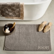 Graue Badematte mit 'JOOP!' Schriftzug, platziert vor einer Badewanne, umgeben von einem Korb mit Pflegeprodukten und beigen Hausschuhen, aus der Vogelperspektive.