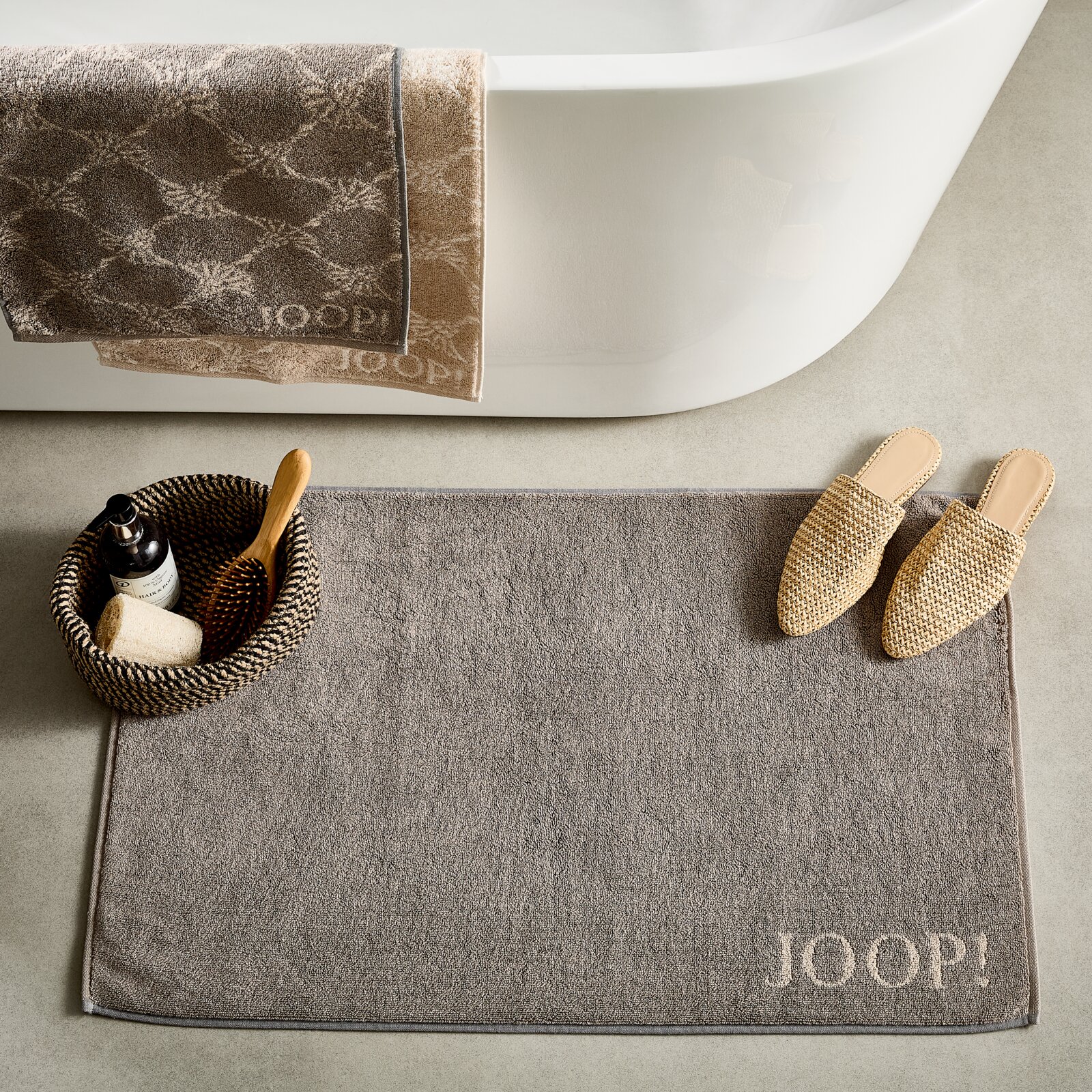 Graue Badematte mit 'JOOP!' Schriftzug, platziert vor einer Badewanne, umgeben von einem Korb mit Pflegeprodukten und beigen Hausschuhen, aus der Vogelperspektive.