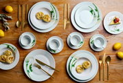 Draufsicht auf ein elegantes weißes Porzellan-Tafelservice mit grünem Blumenmotiv, arrangiert auf einem Holztisch mit goldfarbenem Besteck, Tassen, Zitronen und Dessertteller.
