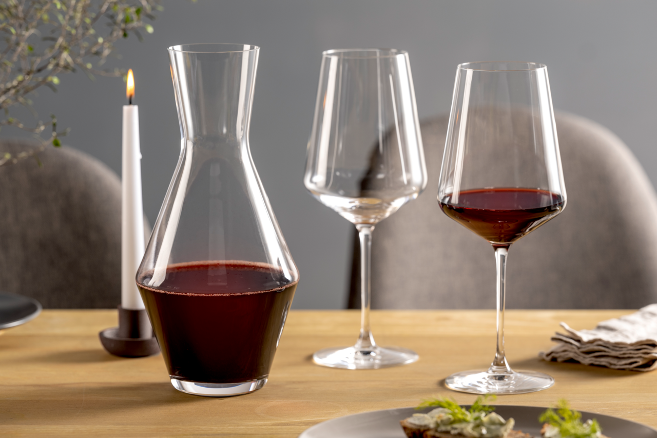 Weinset bestehend aus einer Karaffe und zwei Weingläsern, gefüllt mit Rotwein, auf einem gedeckten Tisch mit Kerze, seitliche Perspektive