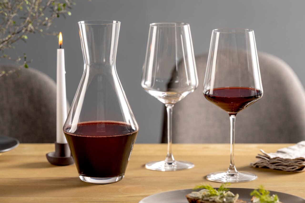 Weinset bestehend aus einer Karaffe und zwei Weingläsern, gefüllt mit Rotwein, auf einem gedeckten Tisch mit Kerze, seitliche Perspektive