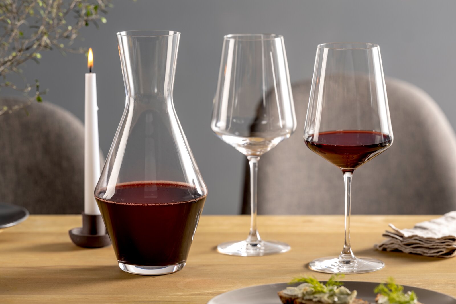 Weinset bestehend aus einer Karaffe und zwei Weingläsern, gefüllt mit Rotwein, auf einem gedeckten Tisch mit Kerze, seitliche Perspektive