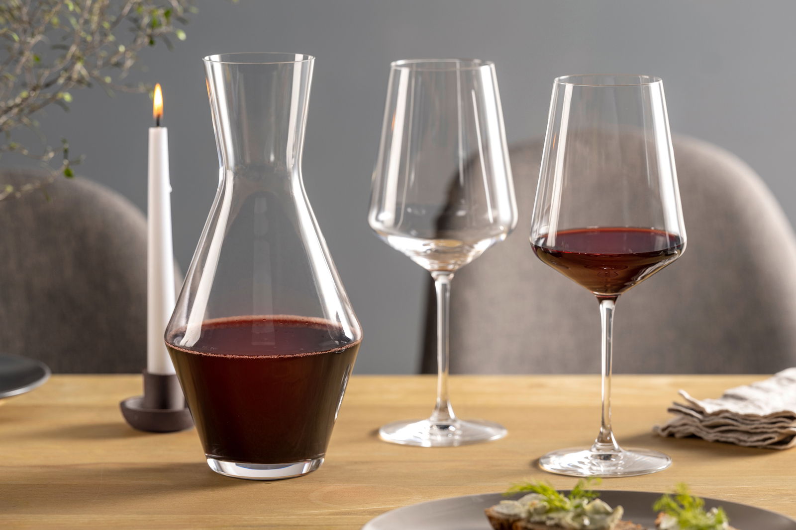 Weinset bestehend aus einer Karaffe und zwei Weingläsern, gefüllt mit Rotwein, auf einem gedeckten Tisch mit Kerze, seitliche Perspektive