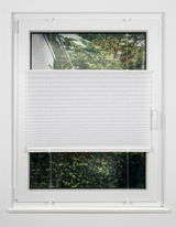 Weiße Plissee-Jalousie an einem Fenster, halb heruntergelassen, mit Blick auf einen Garten. Frontalansicht.