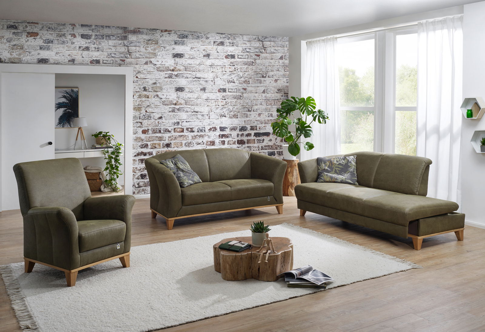 Wohnzimmer mit einem olivgrünen Hochlehnsessel, einem 2,5-Sitzer-Sofa und einer Recamiere mit Armteil links, alle mit sichtbaren Holzsockeln aus Eiche, auf einem hellen Teppich vor einer Backsteinwand. Perspektive: Frontalansicht.