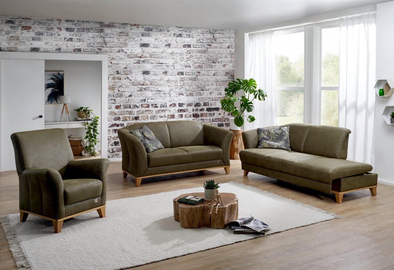 Wohnzimmer mit einem olivgrünen Hochlehnsessel, einem 2,5-Sitzer-Sofa und einer Recamiere mit Armteil links, alle mit sichtbaren Holzsockeln aus Eiche, auf einem hellen Teppich vor einer Backsteinwand. Perspektive: Frontalansicht.