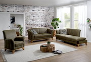 Wohnzimmer mit einem olivgrünen Hochlehnsessel, einem 2,5-Sitzer-Sofa und einer Recamiere mit Armteil links, alle mit sichtbaren Holzsockeln aus Eiche, auf einem hellen Teppich vor einer Backsteinwand. Perspektive: Frontalansicht.