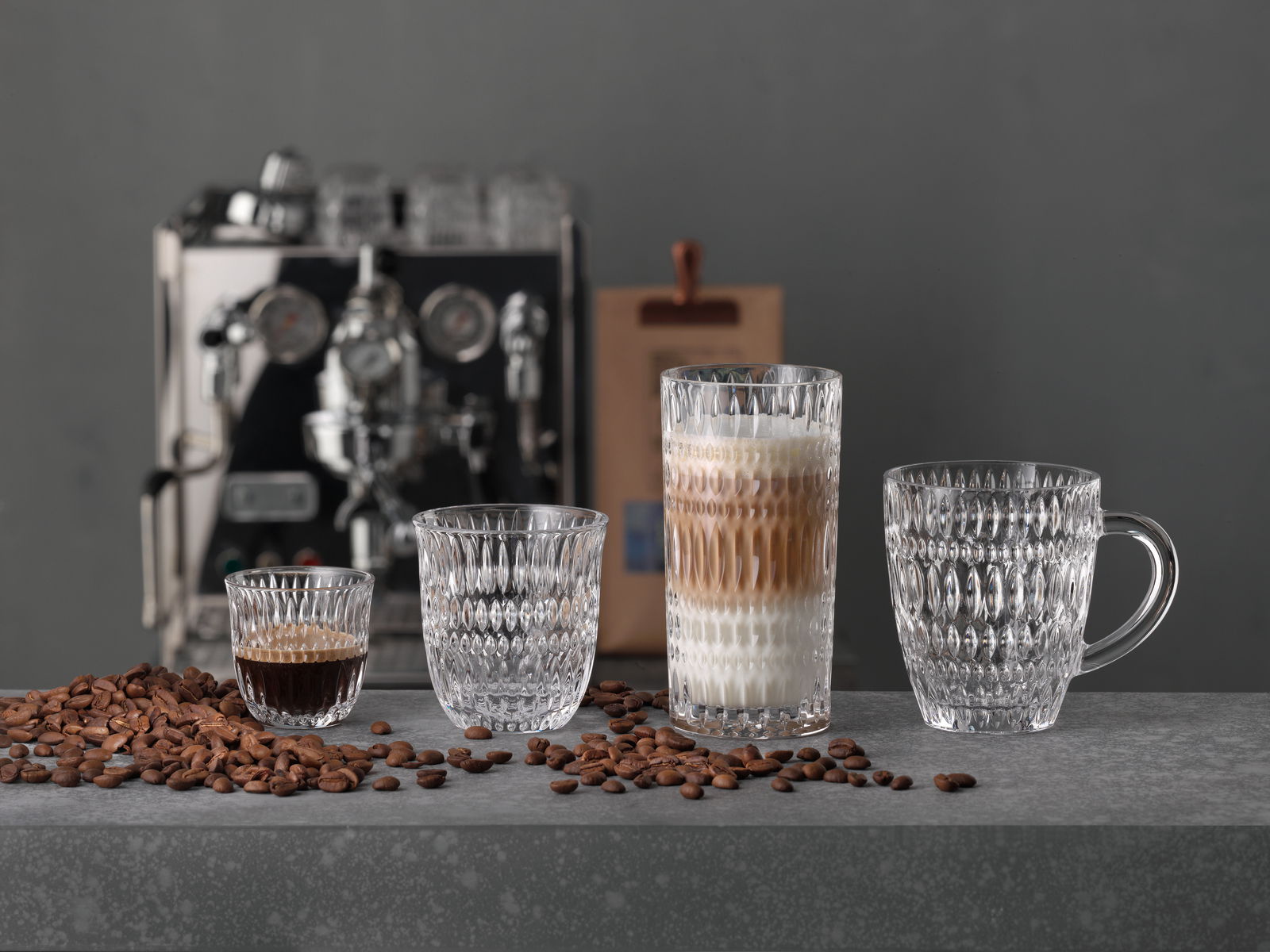 Vier elegante Gläser in verschiedenen Größen für Espresso und Kaffee, auf einer grauen Oberfläche mit Kaffeebohnen, im Hintergrund eine Espressomaschine. Frontalansicht.