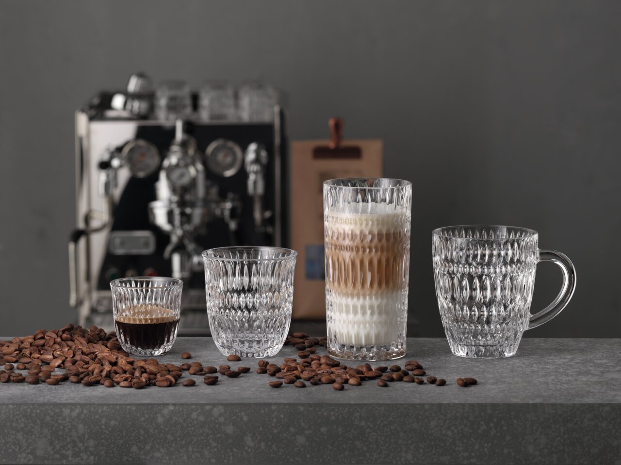 Vier elegante Gläser in verschiedenen Größen für Espresso und Kaffee, auf einer grauen Oberfläche mit Kaffeebohnen, im Hintergrund eine Espressomaschine. Frontalansicht.