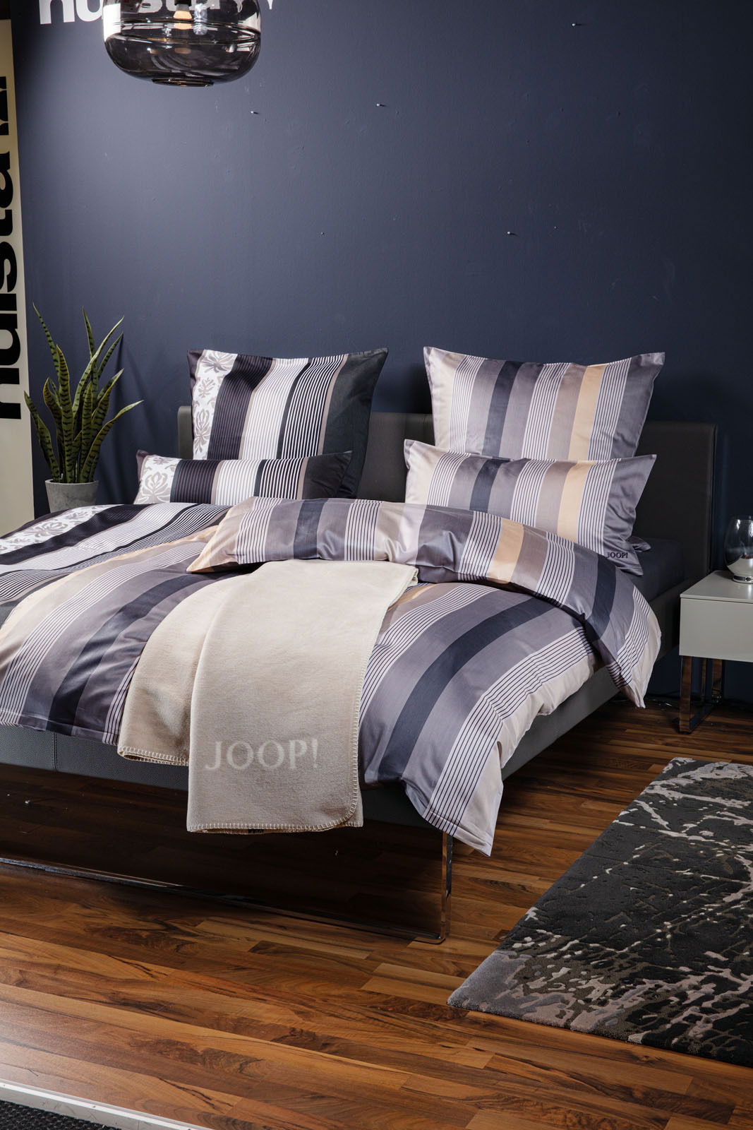 JOOP! Kissenbezug ORNAMENT STRIPE EX Bett mit gestreifter Bettwäsche in Blau- und Beigetönen, dekoriert mit Kissen und einer beigen Decke mit 'JOOP!' Schriftzug, seitliche Perspektive.