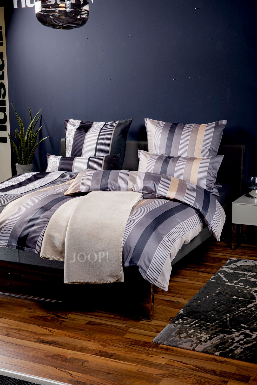 JOOP! Kissenbezug ORNAMENT STRIPE EX Bett mit gestreifter Bettwäsche in Blau- und Beigetönen, dekoriert mit Kissen und einer beigen Decke mit 'JOOP!' Schriftzug, seitliche Perspektive.