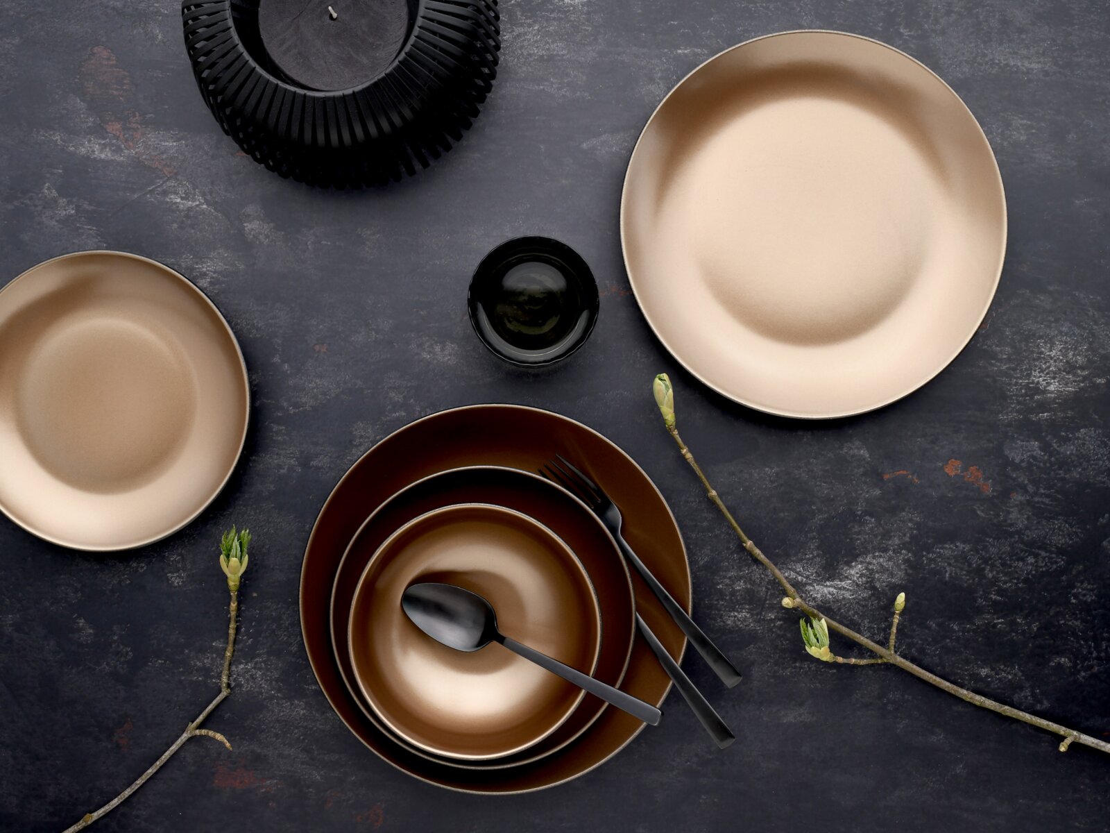 Draufsicht auf ein modernes Tischgedeck mit beige- und braunfarbenen Keramik-Tellern und Schalen, schwarzem Besteck und kleinem schwarzen Schälchen auf dunkler Steinoptik-Oberfläche, dekoriert mit Zweigen.