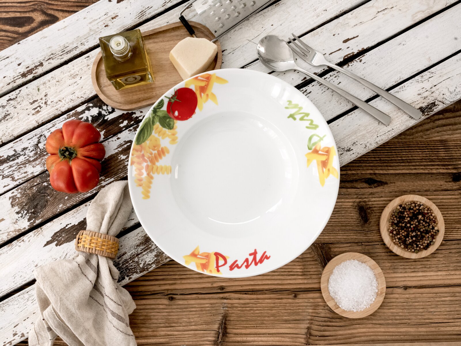 Runder weißer Pastateller mit farbigem Pasta- und Tomatenmotiv und Schriftzug „Pasta“, von oben (Top-Down) auf rustikalem Holztisch arrangiert mit Besteck, Tomate, Ölflasche, Käse, Reibe sowie Schalen mit Salz und Pfeffer.