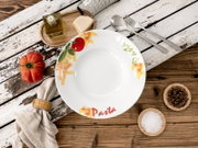 Runder weißer Pastateller mit farbigem Pasta- und Tomatenmotiv und Schriftzug „Pasta“, von oben (Top-Down) auf rustikalem Holztisch arrangiert mit Besteck, Tomate, Ölflasche, Käse, Reibe sowie Schalen mit Salz und Pfeffer.