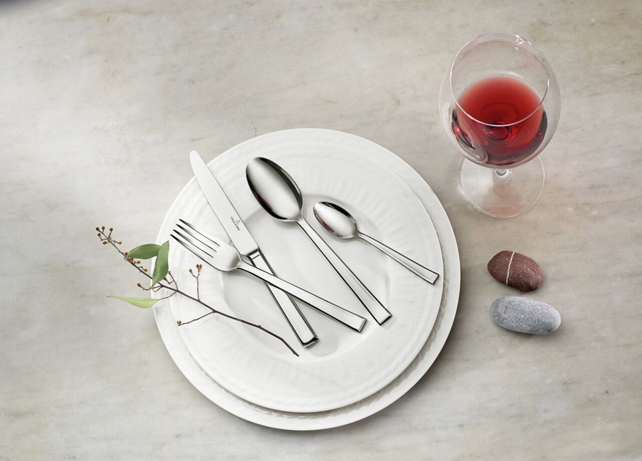 Villeroy & Boch Tafelbesteck 68-tlg. VICTOR Elegantes Tafelbesteck-Set auf einem weißen Teller, mit einem Weinglas und dekorativen Steinen, aus der Vogelperspektive fotografiert.