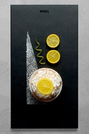 Vogelperspektive auf ein stilvolles Frühstücksset mit einer kleinen Torte, dekoriert mit Zitronenscheiben und Puderzucker, auf einer schwarzen Platte mit dem Logo 'WOLL'.