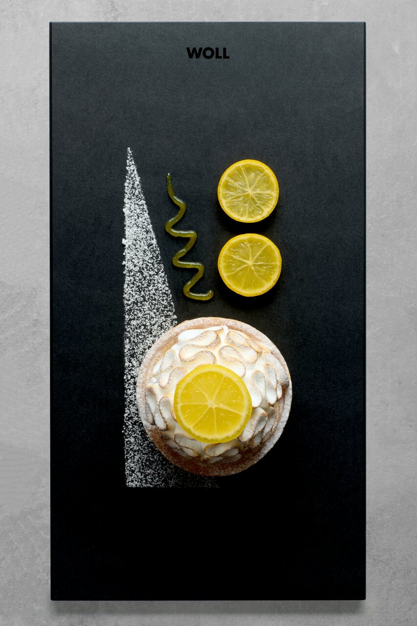 Vogelperspektive auf ein stilvolles Frühstücksset mit einer kleinen Torte, dekoriert mit Zitronenscheiben und Puderzucker, auf einer schwarzen Platte mit dem Logo 'WOLL'.