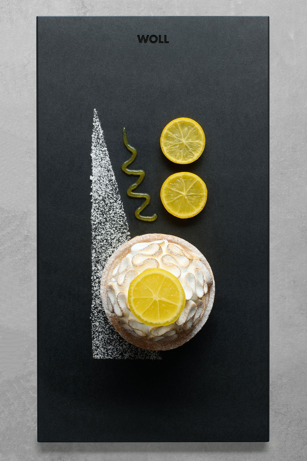 Vogelperspektive auf ein stilvolles Frühstücksset mit einer kleinen Torte, dekoriert mit Zitronenscheiben und Puderzucker, auf einer schwarzen Platte mit dem Logo 'WOLL'.