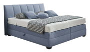 Boxspringbett in blauer Farbe mit gestepptem Kopfteil, seitlicher Perspektive, dekoriert mit gemusterten Kissen und Decke.