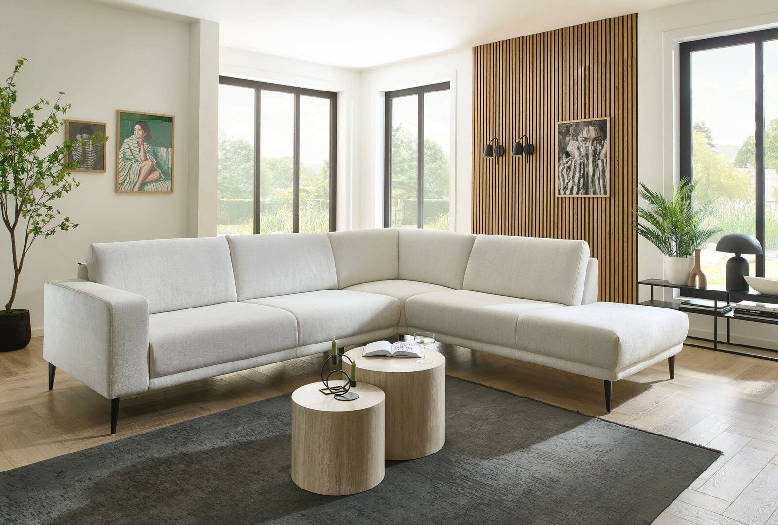 Moderne Eckgarnitur in hellem Stoff, fotografiert aus einer leicht seitlichen Perspektive in einem stilvoll eingerichteten Wohnzimmer mit großen Fenstern und Holzakzenten.