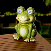 Solarbetriebener Frosch aus Polyresin mit leuchtenden Augen, auf einem Tisch im Freien platziert, frontal fotografiert.