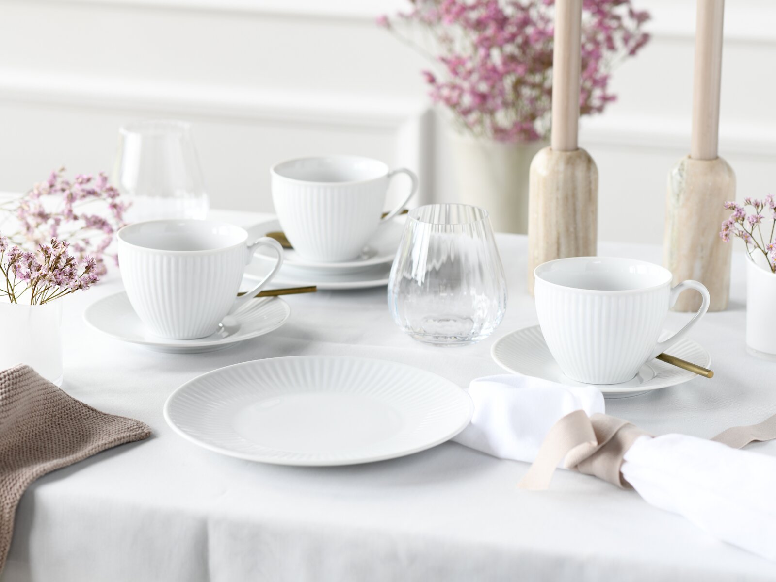 Schlicht gedeckter Tisch mit weißen, geriffelten Kaffeetassen auf Untertassen, weißem Teller, Wassergläsern, goldfarbenen Löffeln und zarten rosa Blumen; Perspektive: leicht erhöhte Frontansicht (schräg von vorne)