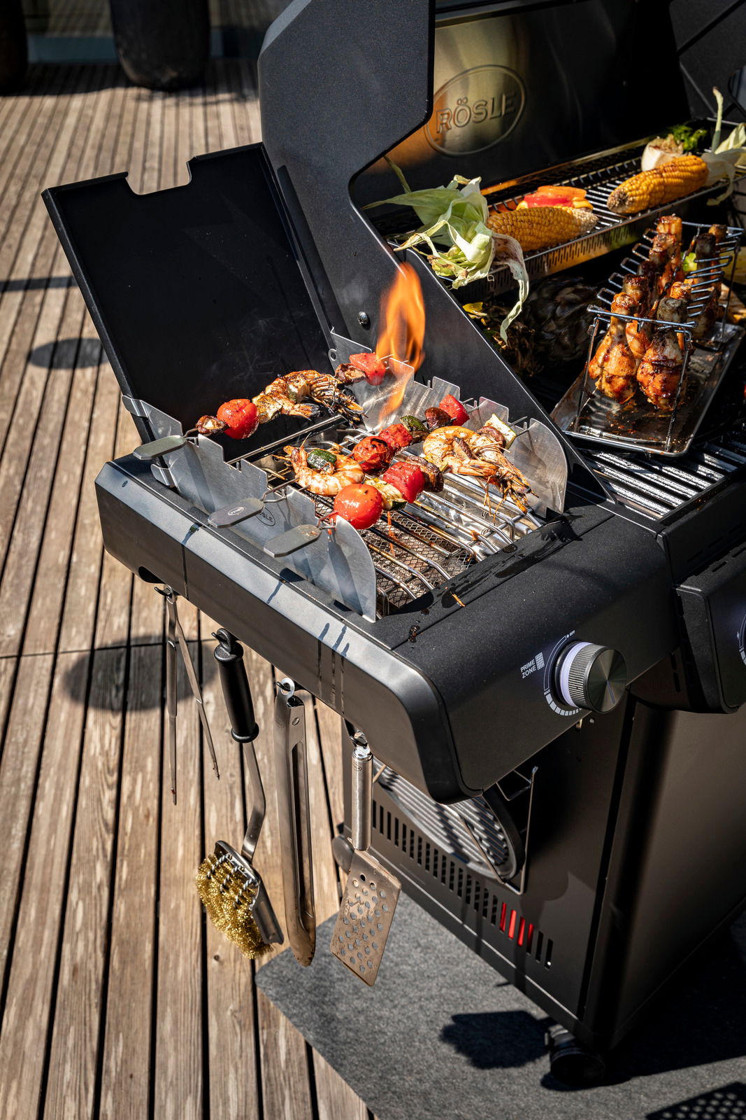 Gasgrill Allflame Hero 4 in seitlicher Perspektive mit gegrilltem Gemüse und Fleischspießen auf dem Rost, umgeben von Grillzubehör.
