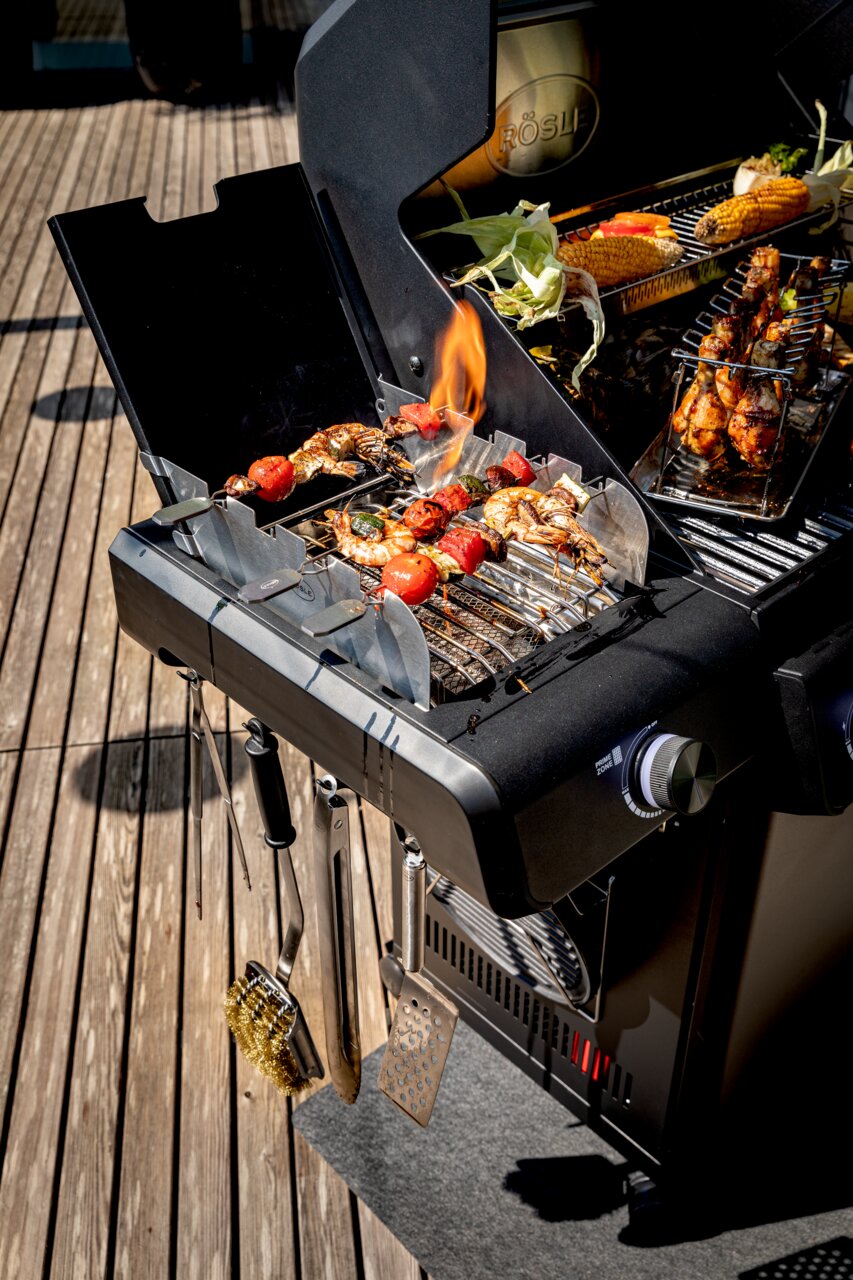 Gasgrill Allflame Hero 4 in seitlicher Perspektive mit gegrilltem Gemüse und Fleischspießen auf dem Rost, umgeben von Grillzubehör.
