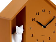 Close-up van een oranje wandklok met een zwarte wijzerplaat en wijzers, en een wit kattenbeeldje in een vakje aan de zijkant.