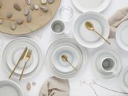 Vogelperspektive auf ein elegantes 16-teiliges Kombiservice 'Ice' mit goldfarbenem Besteck, bestehend aus Tellern, Tassen und Schalen auf einem weißen Hintergrund, dekoriert mit Steinen und Zweigen.