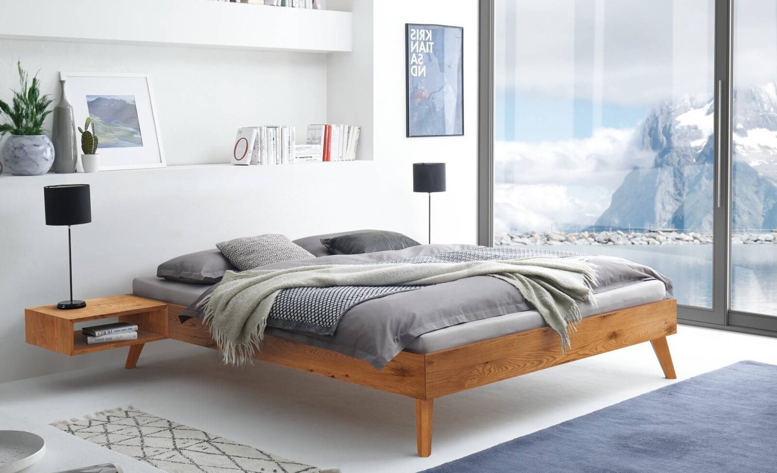 Modern houten bed met grijs beddengoed en een deken in een lichte slaapkamer, gezien vanuit de zijkant (licht schuin van voren) met uitzicht op een grote raampartij en een berglandschap.