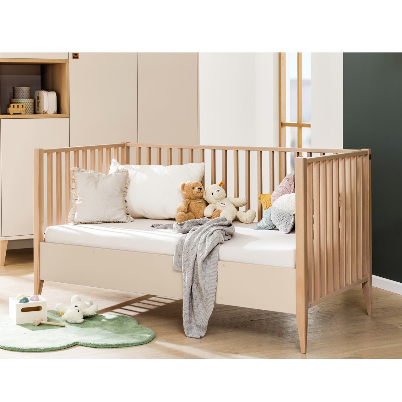 Kinderbett mit Holzgittern, umgeben von Kissen und Kuscheltieren, seitliche Perspektive