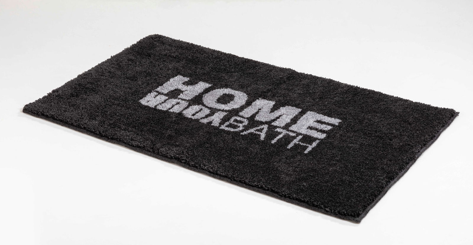 Schwarzer Badteppich mit der Aufschrift 'HOME BATH', perspektivisch von der Seite fotografiert.