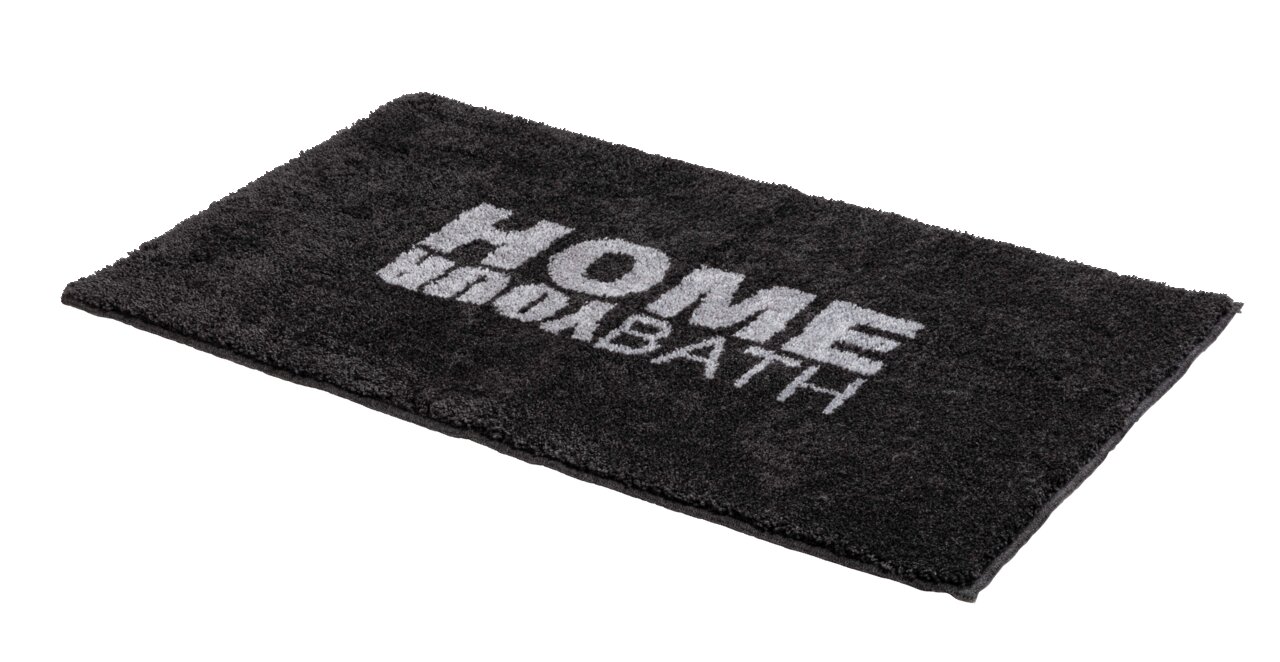 Schwarzer Badteppich mit der Aufschrift 'HOME BATH', perspektivisch von der Seite fotografiert.