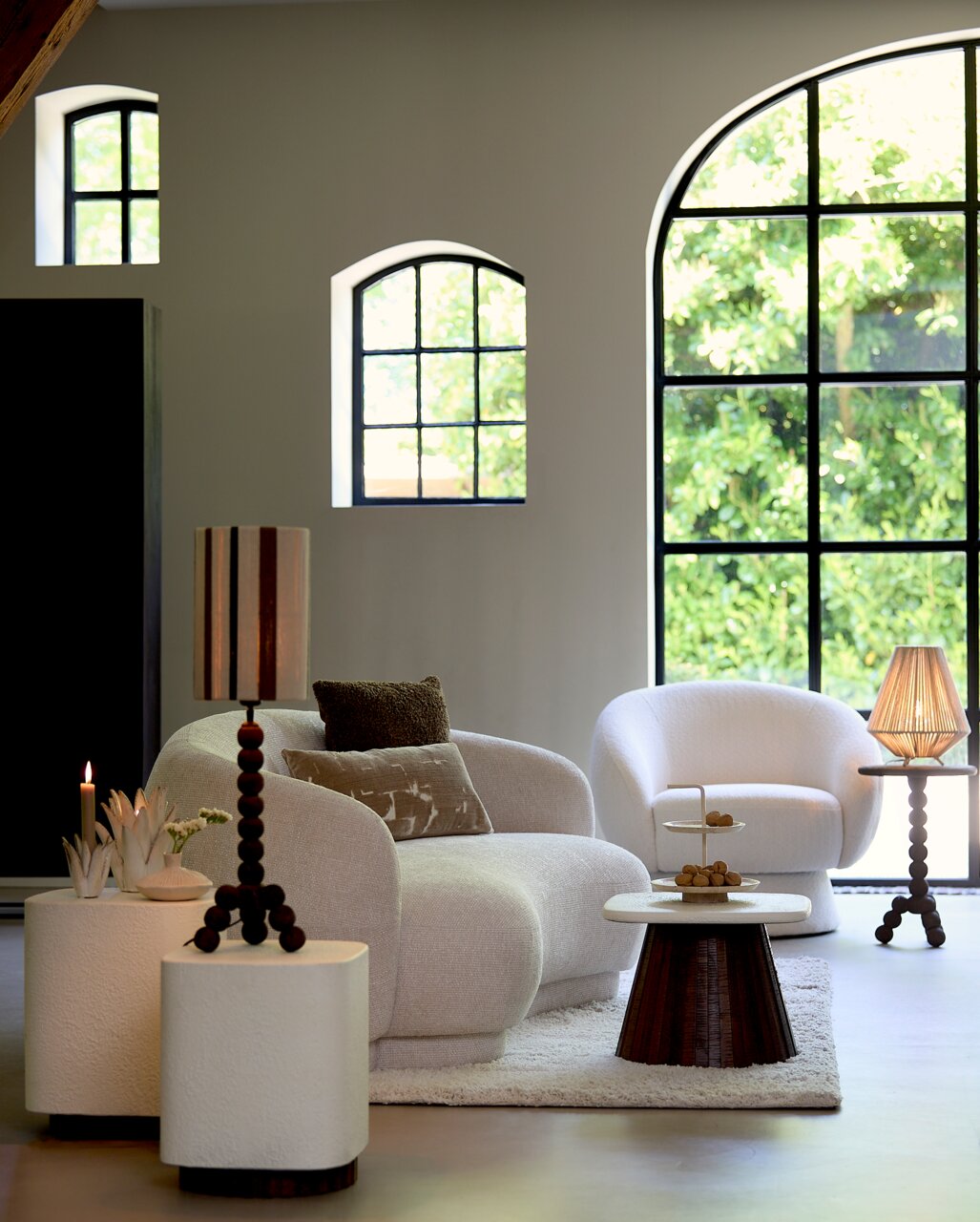 Een gezellig interieur met een elegante, crèmekleurige bank en een bijpassende fauteuil, omringd door stijlvolle bijzettafels en decoraties. Het perspectief toont de meubels van voren in een lichte ruimte met grote ramen.