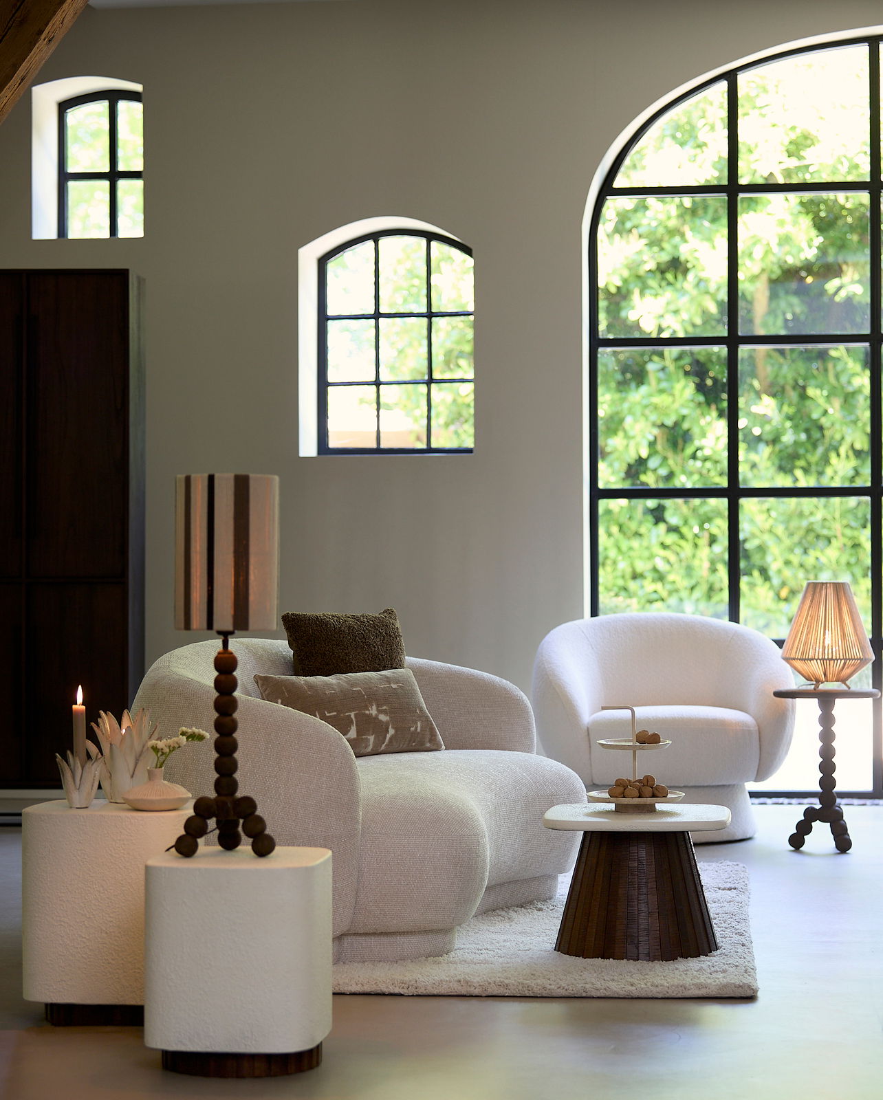 Gemütliche Wohnzimmeransicht mit einer eleganten, cremefarbenen Couch und einem passenden Sessel, umgeben von stilvollen Beistelltischen und Dekorationen. Die Perspektive zeigt die Möbel von vorne in einem hellen Raum mit großen Fenstern.