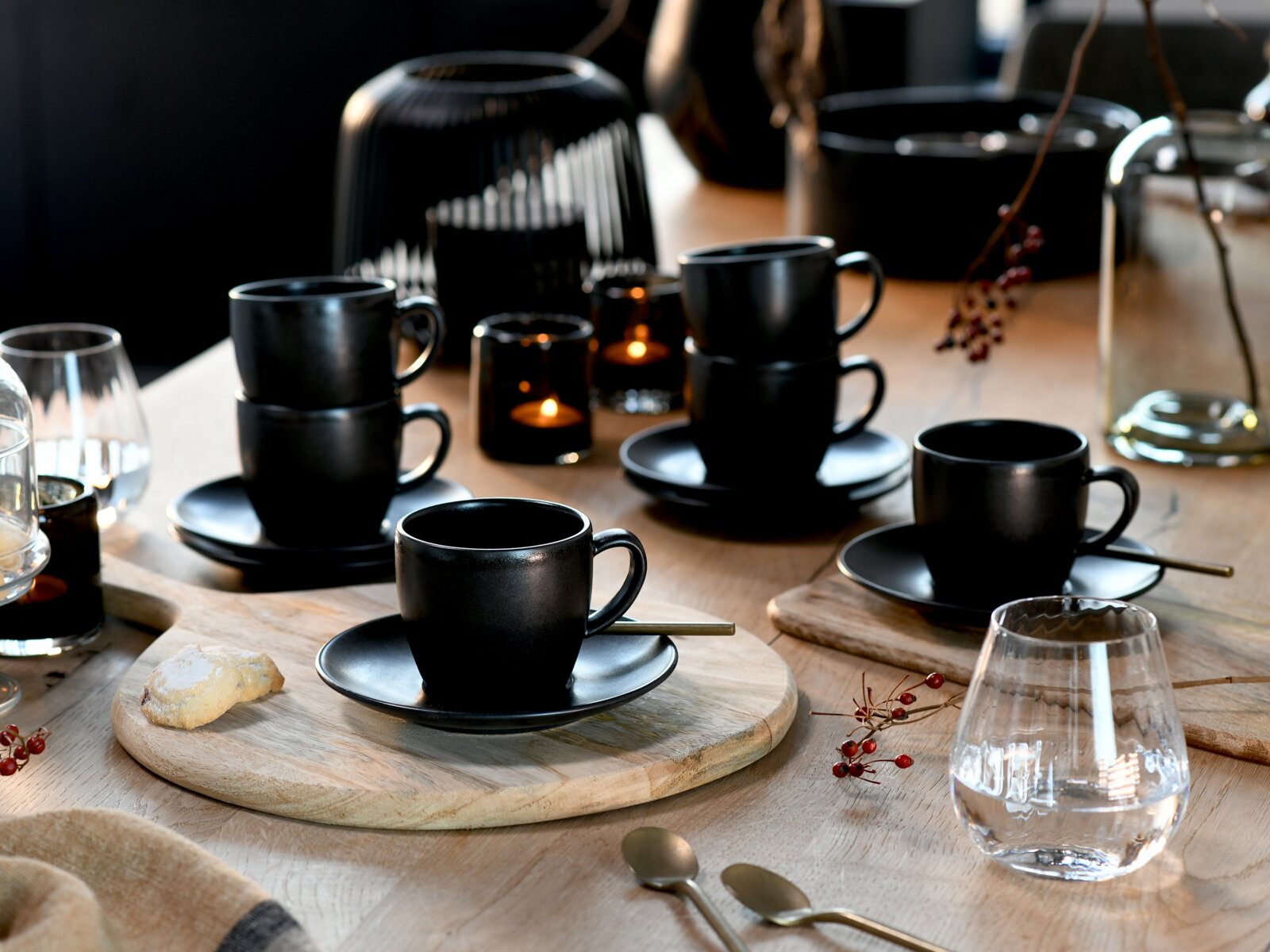 Schwarzes Kaffee- und Espressotassen-Set mit Untertassen auf gedecktem Holztisch, mit Kerzenlicht und Gläsern, fotografiert aus leicht erhöhter schräger Perspektive (Draufsicht).