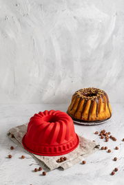 Rote Silikon-Gugelhupfform mit einem gebackenen Gugelhupf im Hintergrund, aus seitlicher Perspektive fotografiert.