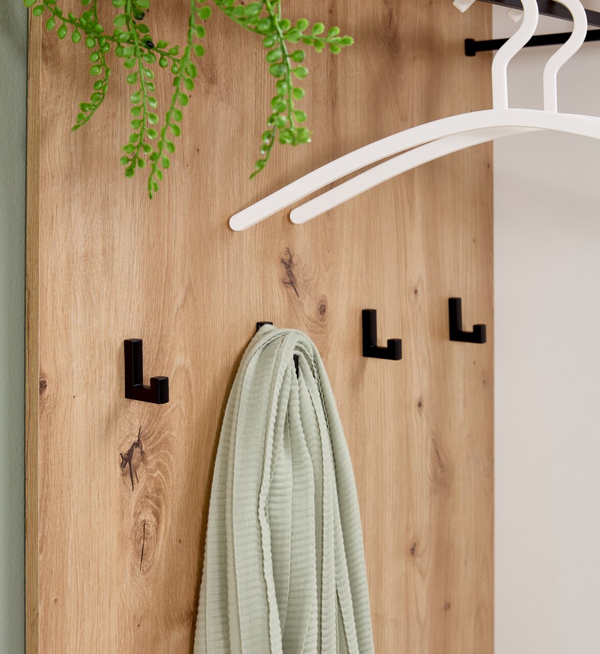 Nahaufnahme einer Kompaktgarderobe aus Holz mit schwarzen Haken, einem grünen Tuch und weißen Kleiderbügeln, von vorne betrachtet.