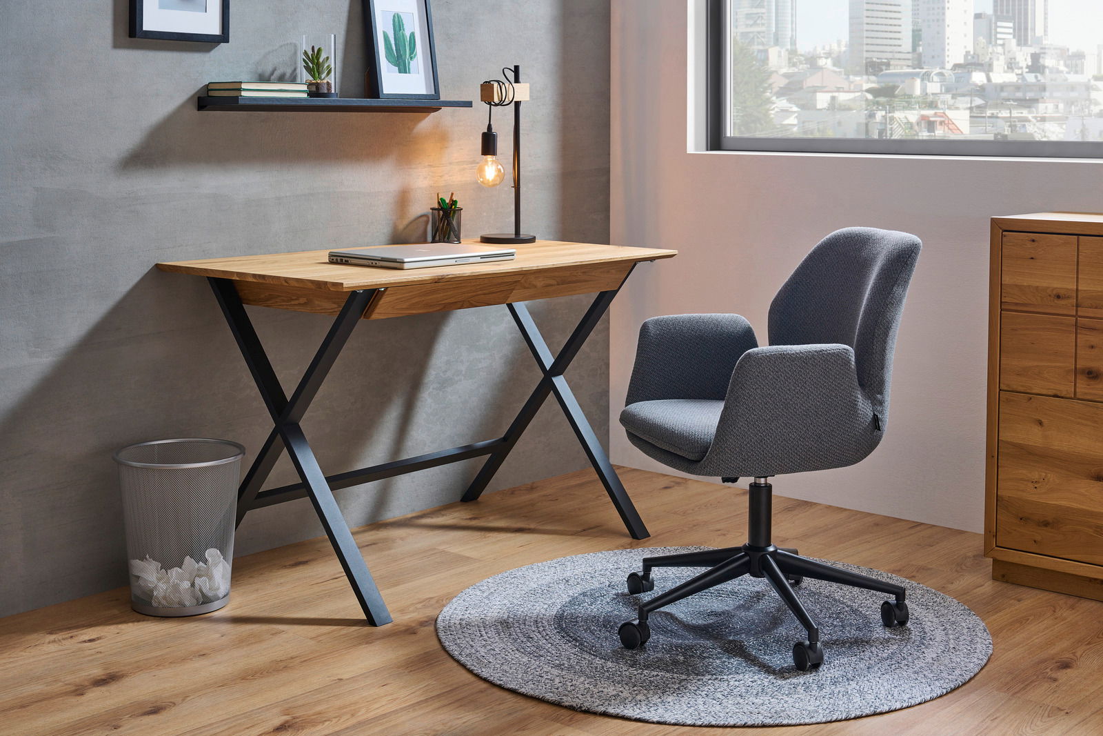 Moderner Schreibtisch mit Holzplatte und schwarzen Metallbeinen, seitliche Perspektive, neben einem grauen Bürostuhl auf einem runden Teppich.