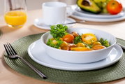 Weiße Schale mit frischem gemischtem Salat (Tomaten, Paprika, Kräuter) auf weißem Teller und grünem Platzset, seitliche Nahaufnahme in leicht schräger Perspektive von vorne; im Hintergrund Tasse und Orangensaft.