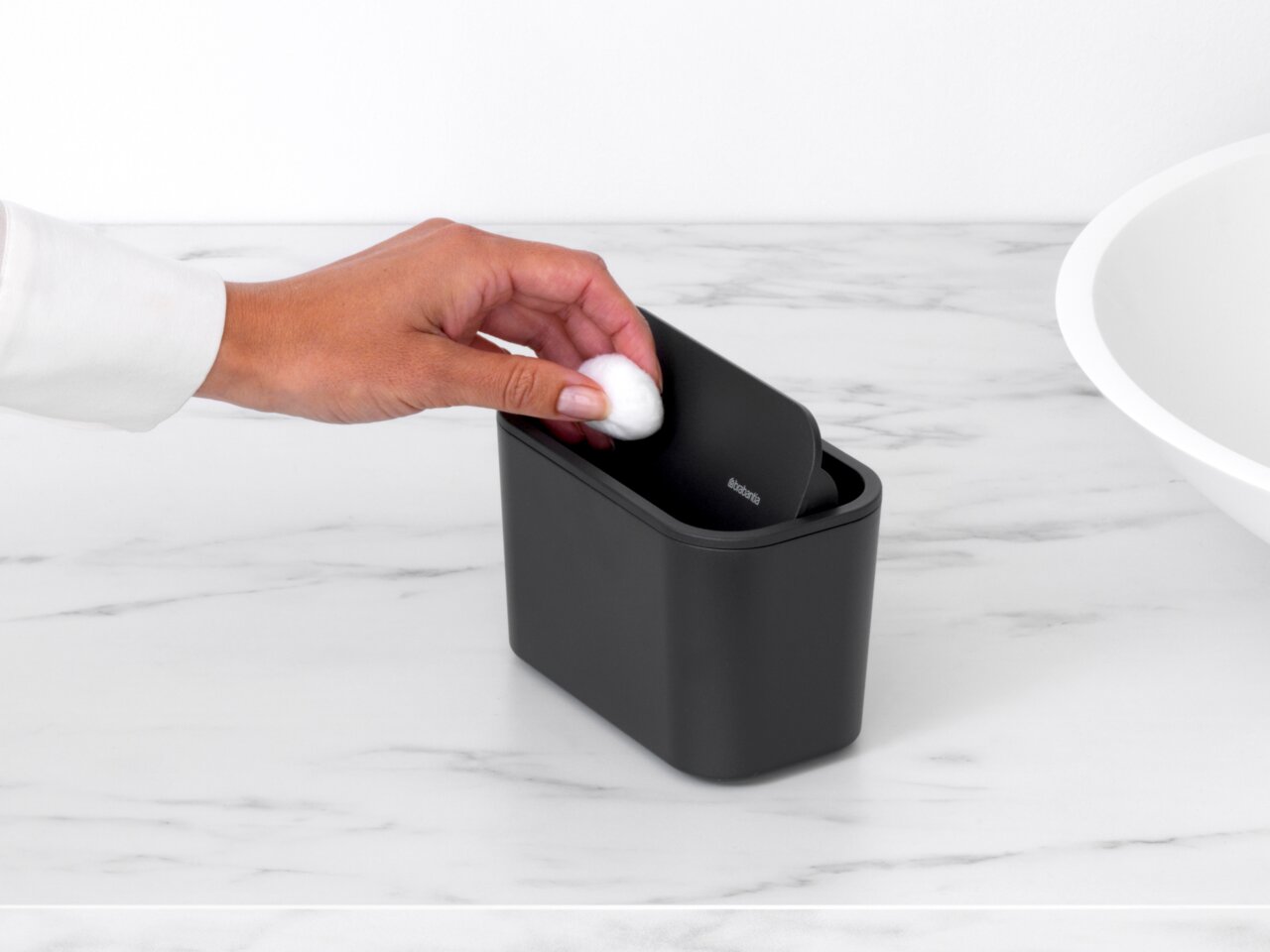 Schwarzer Abfallbehälter auf einer Marmoroberfläche, von oben betrachtet, mit einer Hand, die einen Wattebausch hineinwirft.