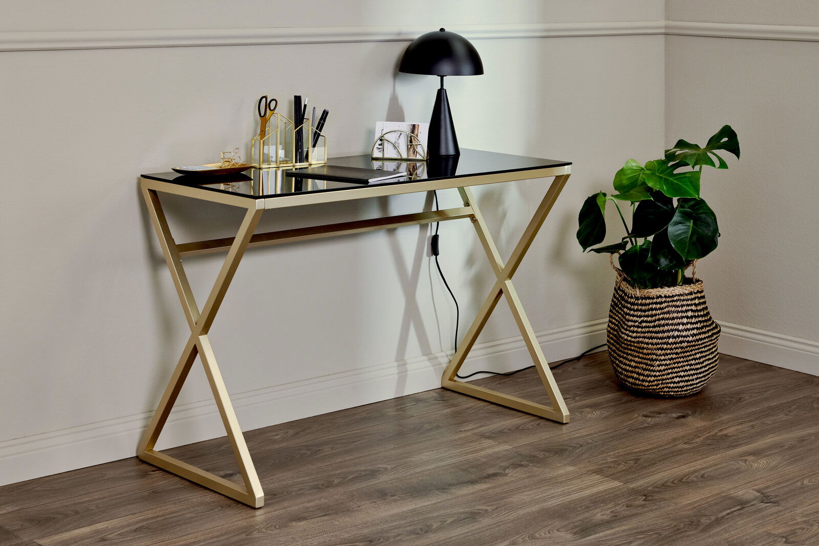 Jahnke Schreibtisch X-TRA D 140 Moderner Schreibtisch mit goldfarbenem Gestell und schwarzer Glasplatte, seitliche Perspektive, dekoriert mit Schreibtischutensilien und einer Lampe, daneben eine Pflanze in einem Korb.