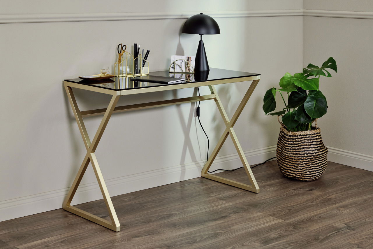 Moderner Schreibtisch mit goldfarbenem Gestell und schwarzer Glasplatte, seitliche Perspektive, dekoriert mit Schreibtischutensilien und einer Lampe, daneben eine Pflanze in einem Korb.