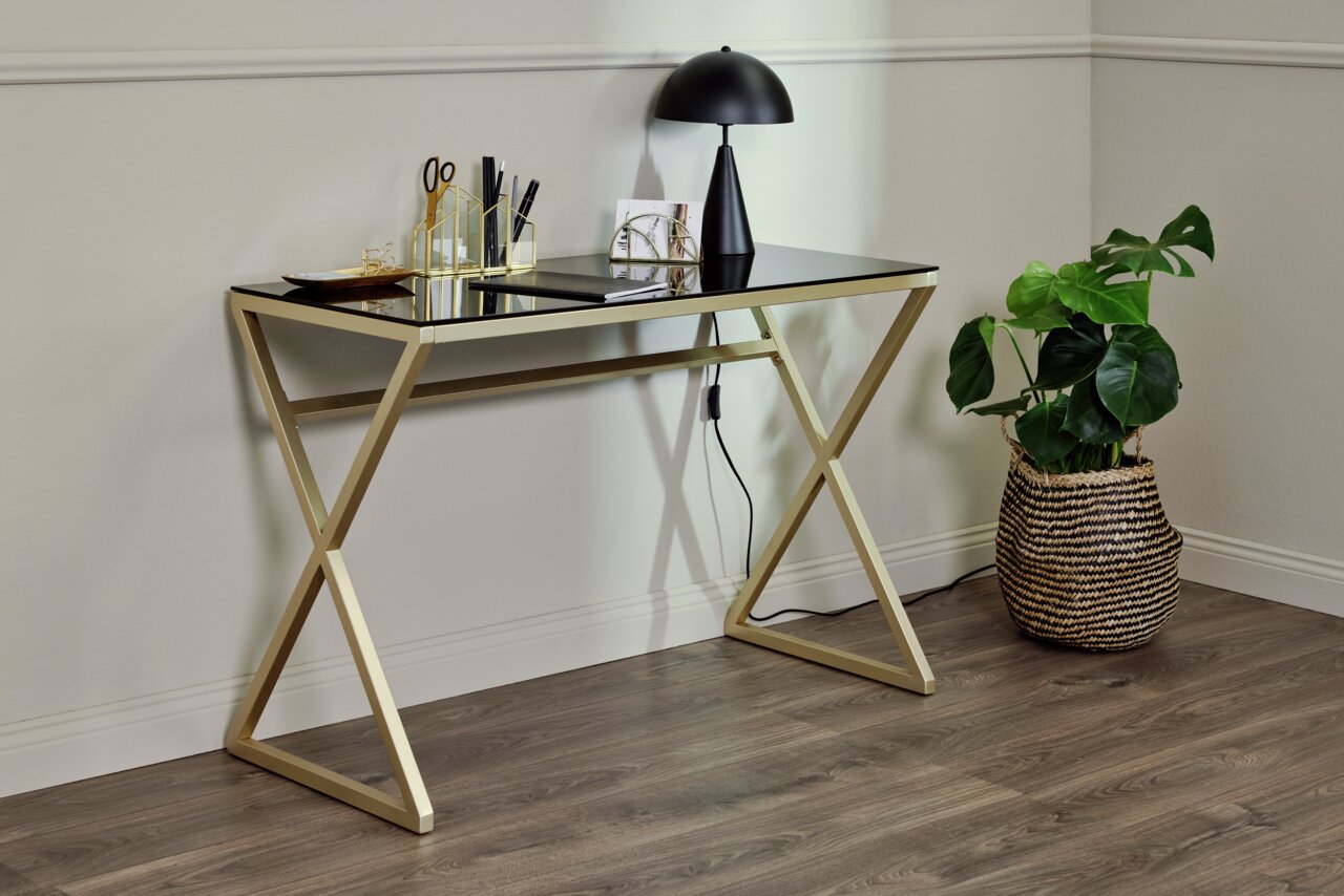Moderner Schreibtisch mit goldfarbenem Gestell und schwarzer Glasplatte, seitliche Perspektive, dekoriert mit Schreibtischutensilien und einer Lampe, daneben eine Pflanze in einem Korb.