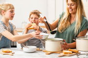 Fleischtopf almond white in einer Küche, von einer Frau mit Suppenkelle bedient, während zwei Kinder Sandwiches essen. Perspektive von vorne.