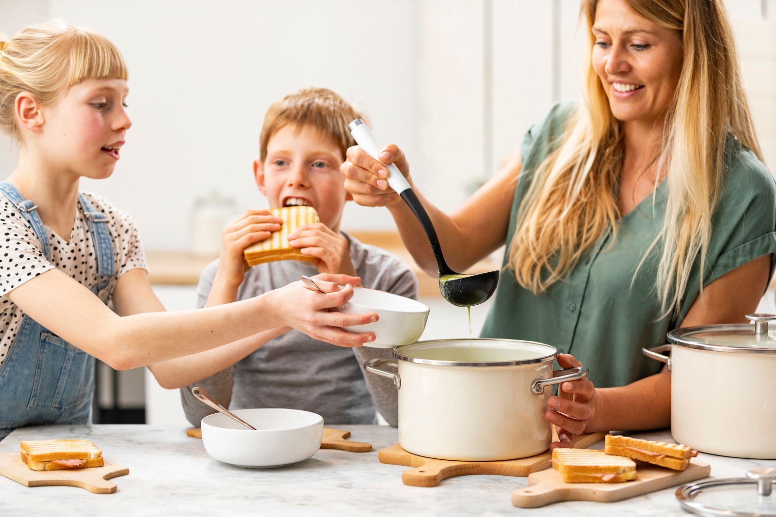 Fleischtopf almond white in einer Küche, von einer Frau mit Suppenkelle bedient, während zwei Kinder Sandwiches essen. Perspektive von vorne.