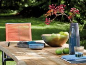 Holz-Esstisch im Freien mit Dekorationen wie einer Schale mit grünen Äpfeln, einer Vase mit roten Beeren und Geschirr, seitliche Perspektive