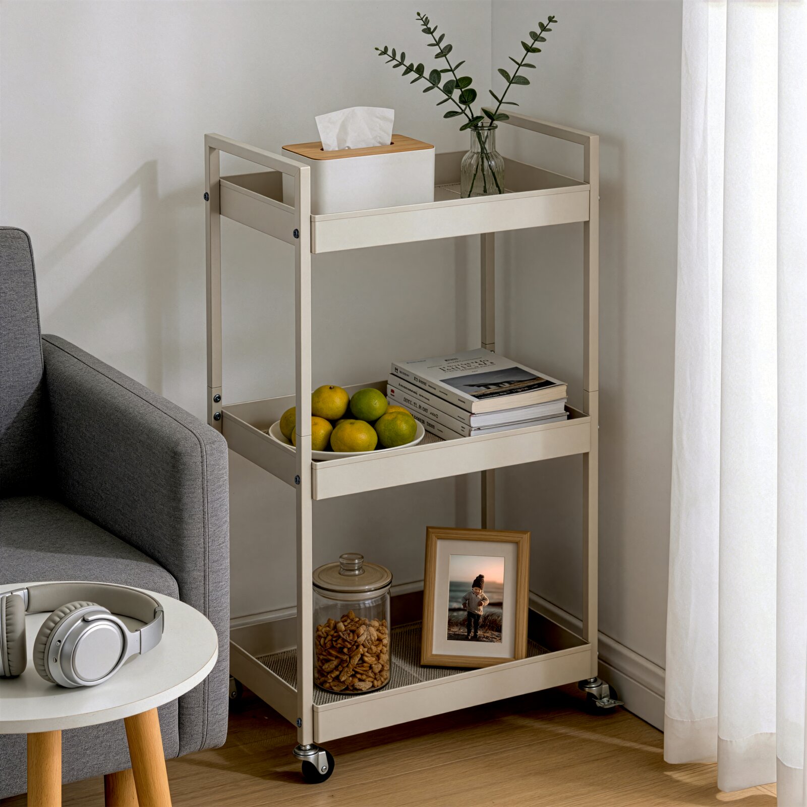 Beiger, dreistufiger Servier- und Aufbewahrungswagen aus Metall auf Rollen neben einem grauen Sofa, mit Taschentuchbox, Glasvase mit Zweigen, Obstschale, Büchern, Keksdose und Bilderrahmen; Ansicht schräg von vorne.